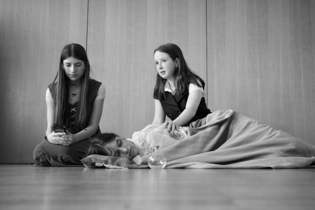 Three girls in a room: one on the left looking at a phone, the middle girl sitting and looking to the side, and a third girl sleeping on a blanket in front.