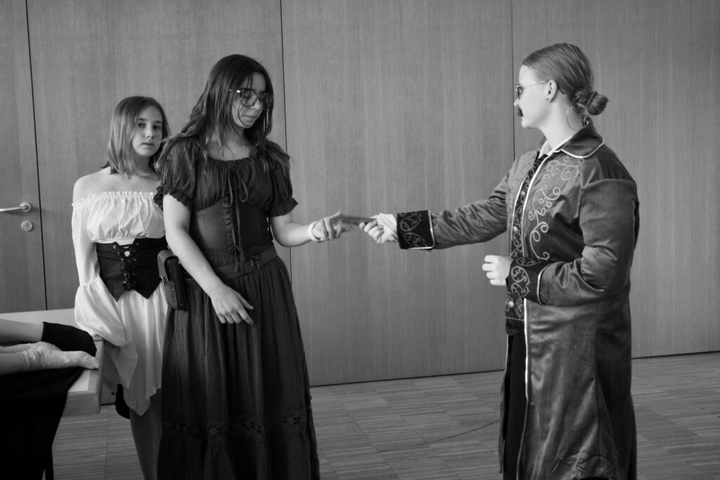 Three people in period costume exchange a document indoors, with a girl in a white off-shoulder blouse observing on the left.